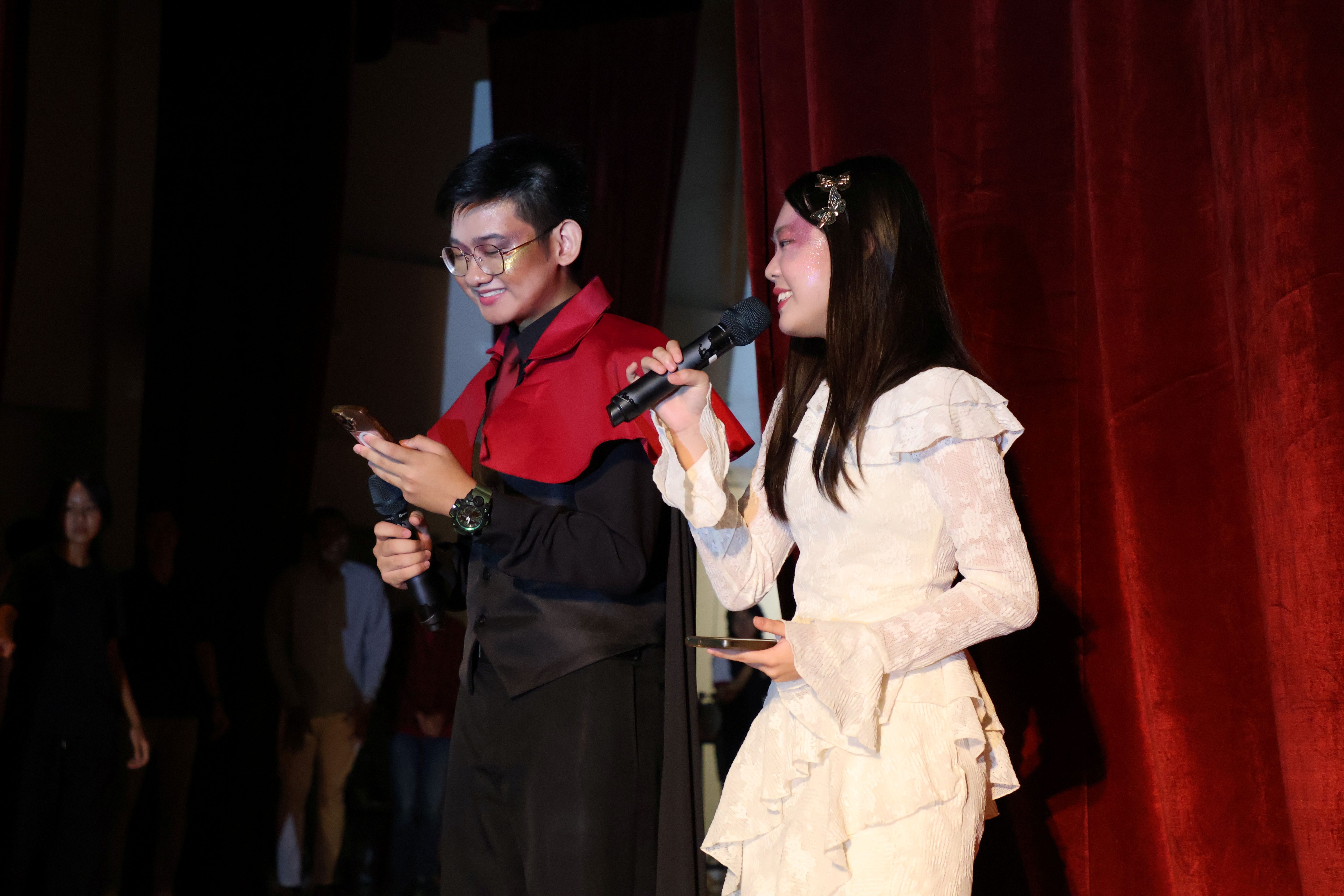 Two student emcees in elaborate costumes hold microphones on stage — one in a dramatic black-and-red cape, the other in a white ruffled dress with glittery makeup — hosting the Teachers' Day celebration.